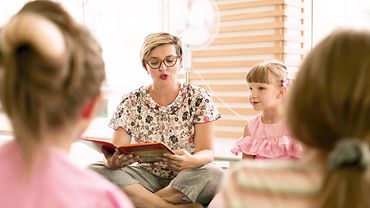Lesezeit in der Kita: Eine Erzieherin liest aus einem Buch, neben ihr sitzt ein Mädchen und hört aufmerksam zu, im Hintergrund ein Ventilator.