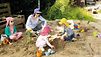 Drei Kinder und eine Erwachsene mit Sonnenbrill und Hut mit Nackenschutz in einem Sandkasten, im Sand liegen Spielgeräte.