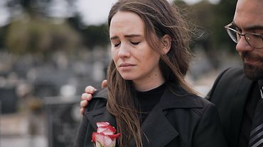 Eine Frau mit traurigem Gesichtsausdruck und geschlossenen Augen in schwarzer Kleidung. Sie hält eine weiß-rote Rose in der Hand. Ein Mann mit Brille legt von hinten eine Hand um ihre Schulter