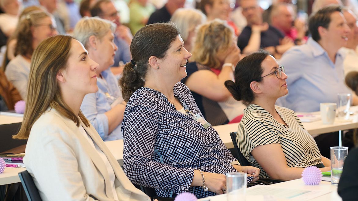 Drei Frauen im Vordergrund sitzen an einem Tisch, lächeln und schauen in eine Richtung, unscharf im hintergrund viele weitere Personen im Publikum an Tischen.