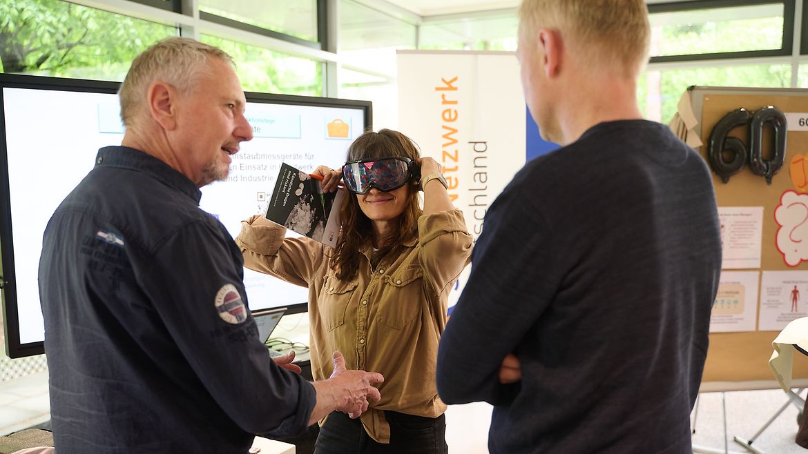 Eine Frau setzt eine VR-Brille auf, während ein ihr gegenüber stehender Mann einem anderen Mann etwas erklärt.