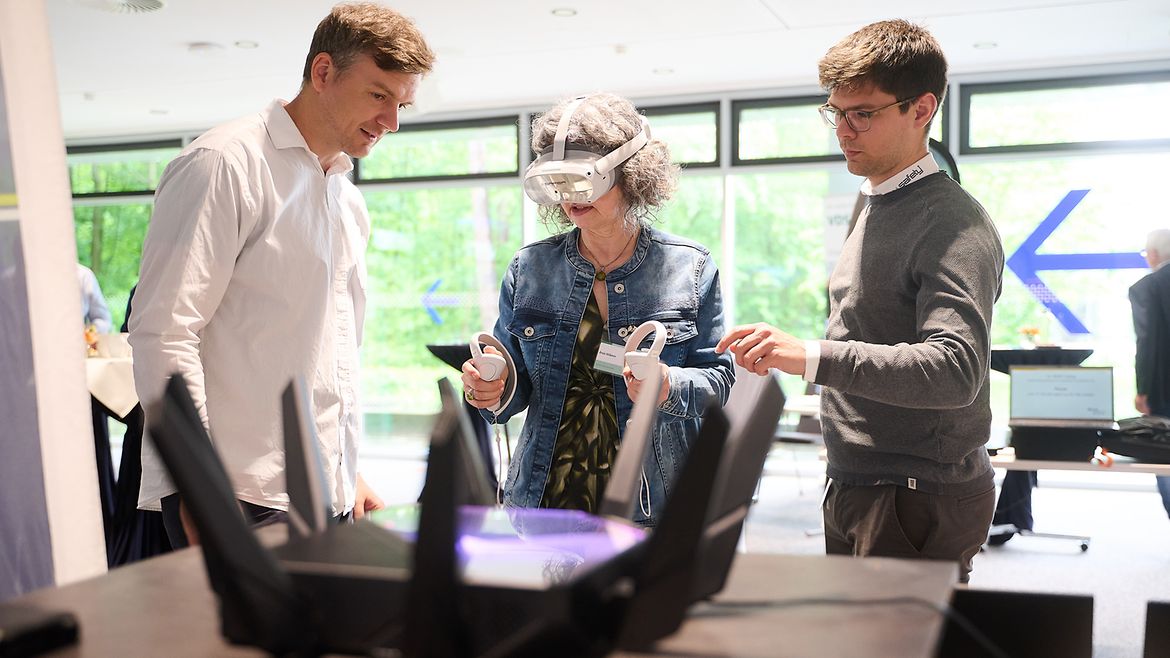 Eine Frau trägt eine VR-Brille auf dem Kopf und Controller in den Händen. Zwei Männer stehen neben ihr.