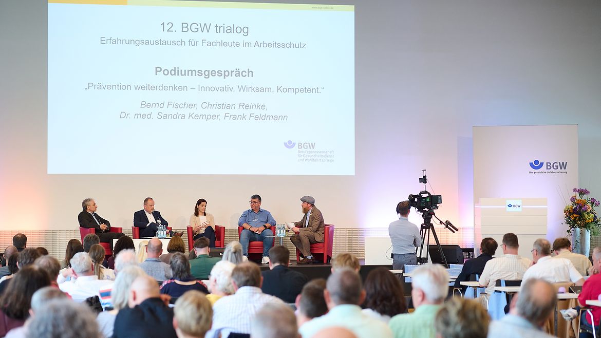 Blick aus dem Publikum auf die Bühne des trialogs: Fünf Personen sitzen in Stühlen zu einem Podiumsgespräch zusammen.