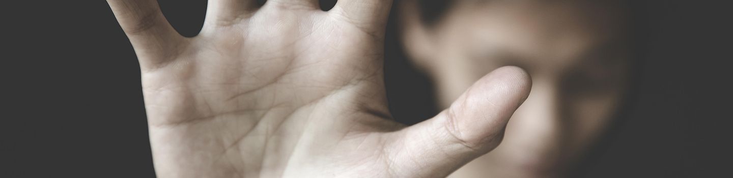 Eine zur Abwehr nach vorn gestreckte Hand im Vordergrund. Hinten verschwommen eine Frau mit gesenktem Kopf.