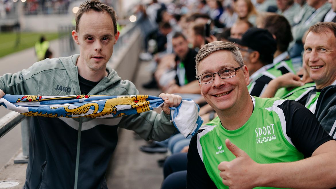 Zwei Teilnehmer bei der Eröffnungsfeier: Einer hält eine Bayern-Flagge, der andere zeigt mit dem Daumen nach oben.