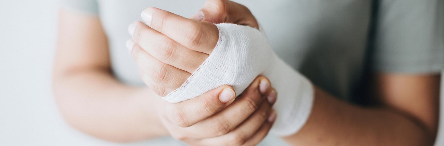 Nahaufnahme der Hände einer Frau. Eine Hand ist mit Mullbinde verbunden. Mit der anderen Hand drückt sie auf die verletzte Stelle