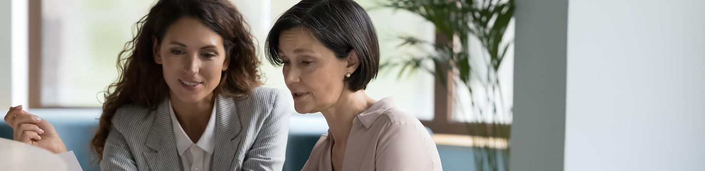 Eine ältere und eine jüngere Frau sitzen gemeinsam vor einem Laptop. Die ältere Frau sitzt rechts und deutet auf den Bildschirm. Sie beraten sich über den Inhalt auf dem Bildschirm.