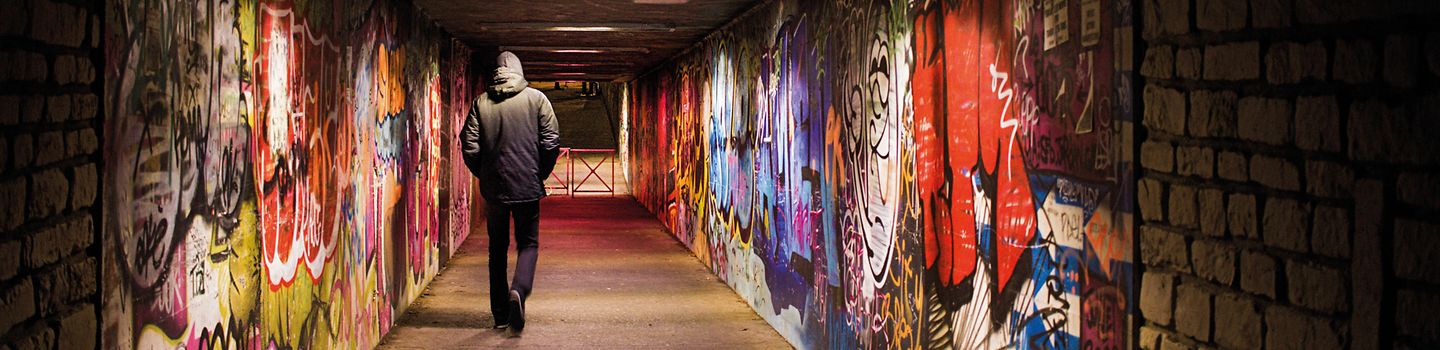 Ein Mann läuft durch eine schwachbeleuchtete Unterführung. Die Wände der Unterführung sind mit Graffitis besprüht.