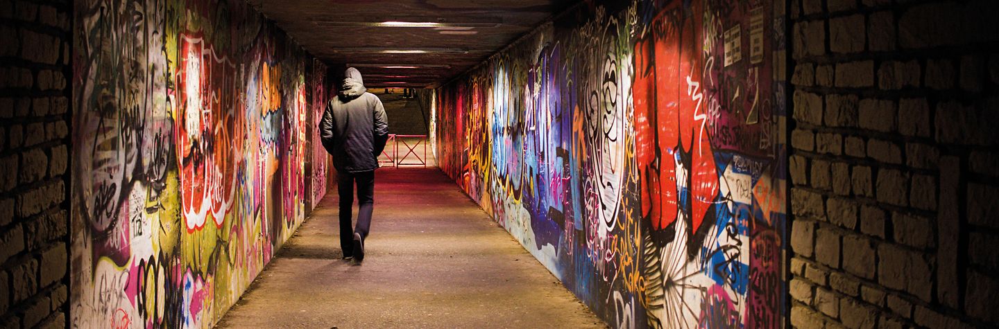 Ein Mann läuft durch eine schwachbeleuchtete Unterführung. Die Wände der Unterführung sind mit Graffitis besprüht.