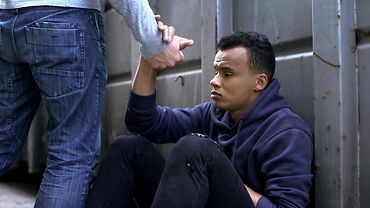 Ein junger Mann in Hoodie und zerrissenen Hosen sitzt mit traurigem Blick vor einer Stahlwand, an die er seinen Rücken lehnt. Die rechte Hand des jungen Manns greift das Handgelenk eines Mannes, der vor ihm steht.