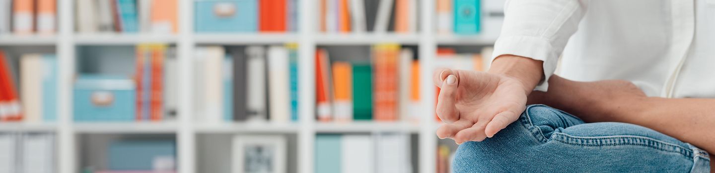 Foto einer Person, die im Schneidersitz auf einem weißen Schreibtisch vor einem Regal sitzt. Das Regal ist mit Büchern und Unterlagen gefüllt.