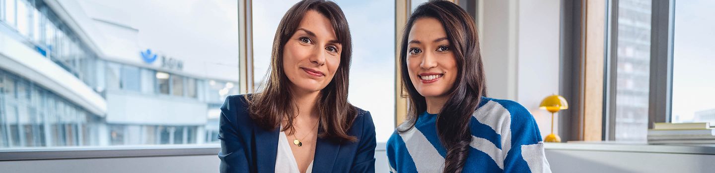 Zwei junge Frauen sitzen nebeneinander vor einem Fenster im Büro und lächeln in die Kamera.