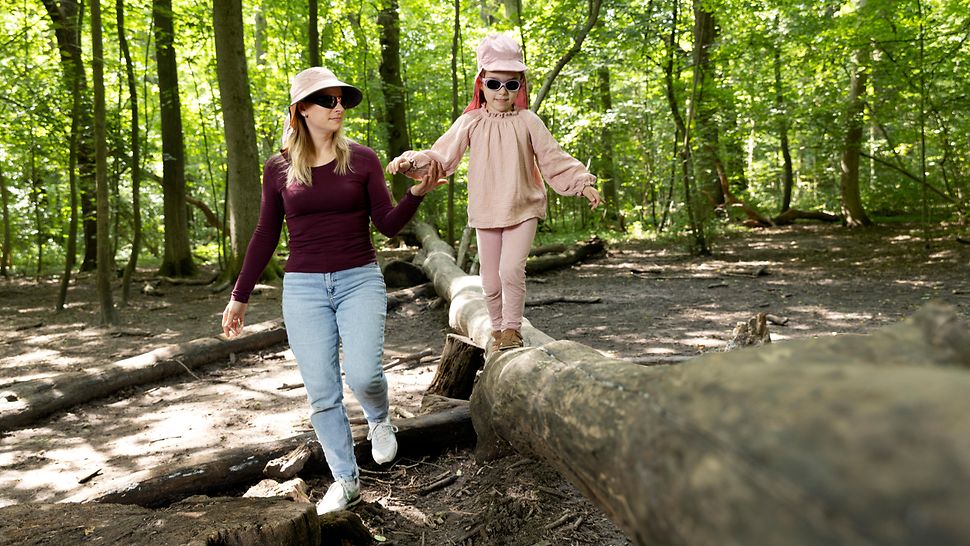 Eine Frau mit Sonnenbrille und Sonnenhut hält die Hand eines Kindes, das im Wald auf einem Baumstamm balanciert.