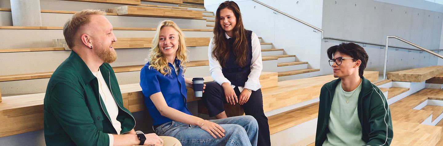 Zwei Männer und zwei Frauen sitzen auf hölzernen Stufen in der Lobby eines Gebäudes, sind im Gespräch und lächeln.