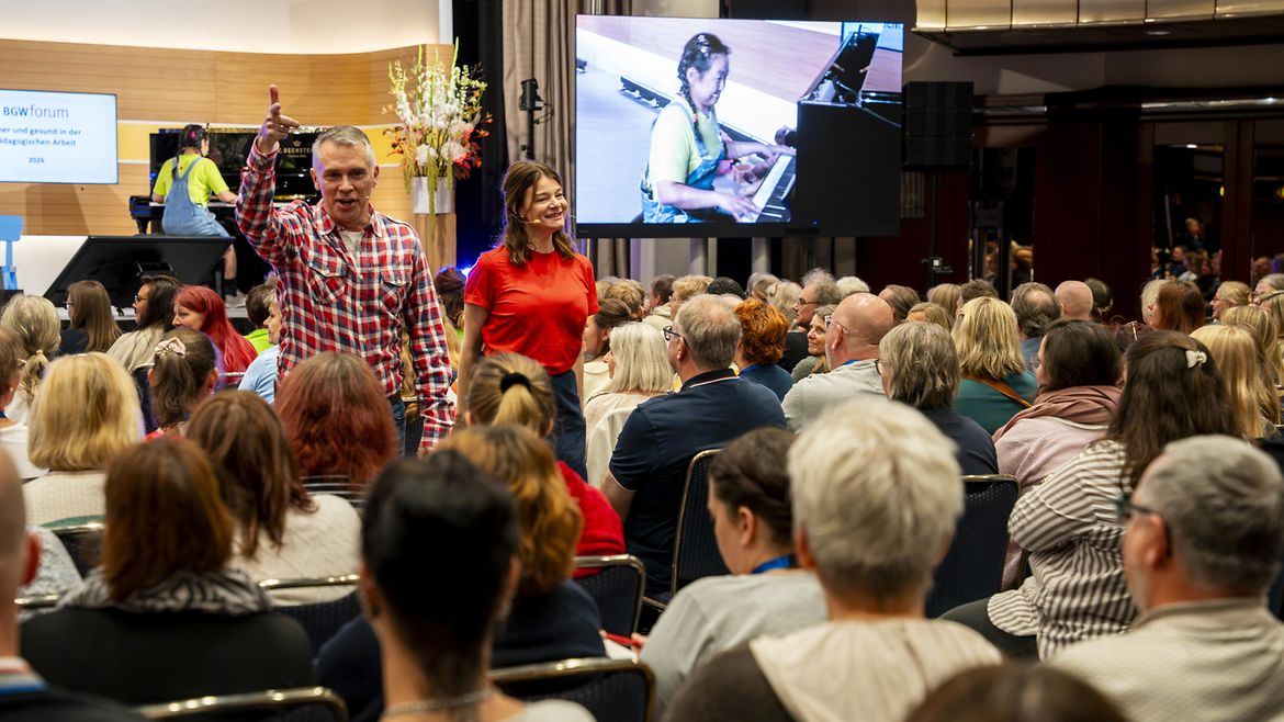 Ein lächelnder Mann und eine lächelnde Frau inmitten eines Publikums, im Hintergrund eine Frau am Klavier.