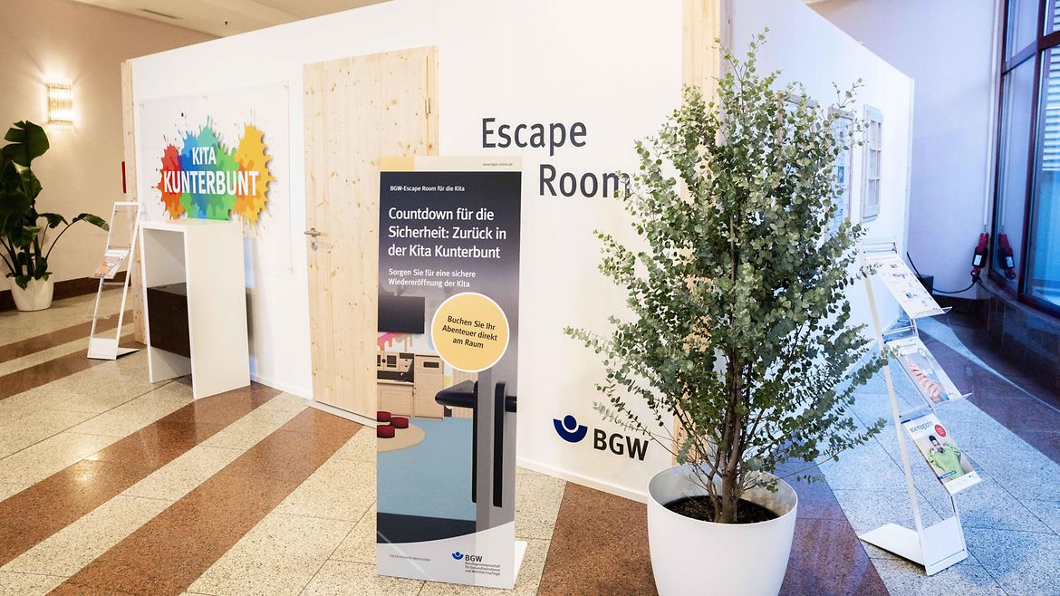 BGW-Messehäuschen mit Holztür, auf der Vorderseite des Häuschens prangen zwei Tafeln. Auf einer steht "Escape Room", auf einer anderen "Kita Kunterbunt". Das Häuschen hat eine Tür und mehrere Sprossenfenster, im Vordergrund ein Aufsteller "Countdown für die Sicherheit: Zurück in der Kita Kunterbunt. Sorgen Sie für eine sichere Wiedereröffnung der Kita. Buchen Sie Ihr Abenteuer direkt am Raum".