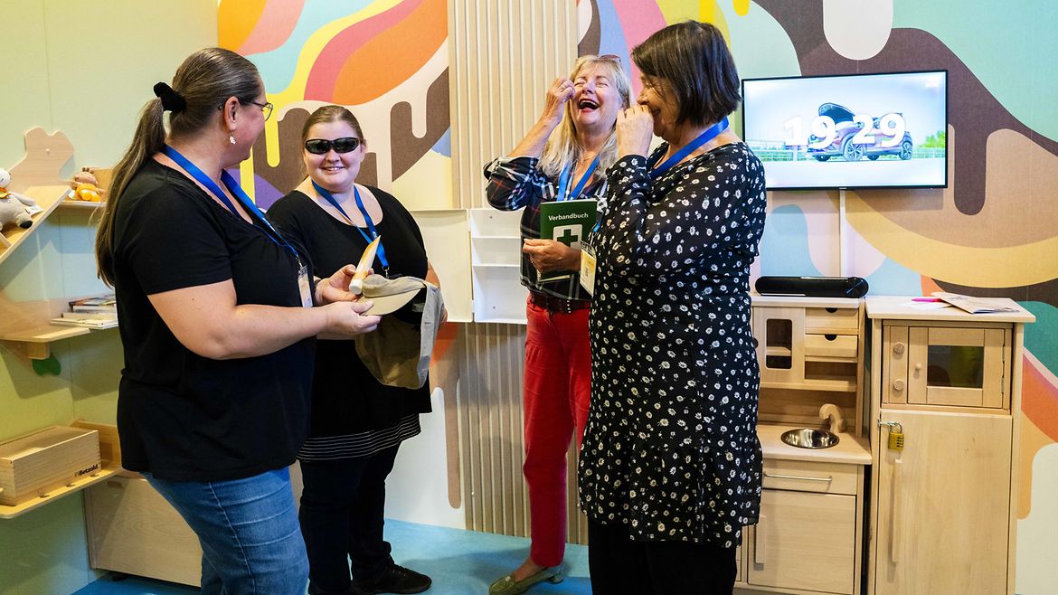 Vier Frauen im Gespräch, alle lachen amüsiert. Die Wand im Hintergrund ist mit bunten Farbklecksen geschmückt, hinten rechts ein hölzernes Regal mit Spielzeug und Büchern, außerdem eine hölzerne Spielküche und ein Monitor.Eine der Frauen trägt eine Sonnenbrille, eine weitere hält eine Tube Sonnencreme sowie einen Hut mit Nackenschut in der Hand, eine weitere Frau ein Verbandbuch.