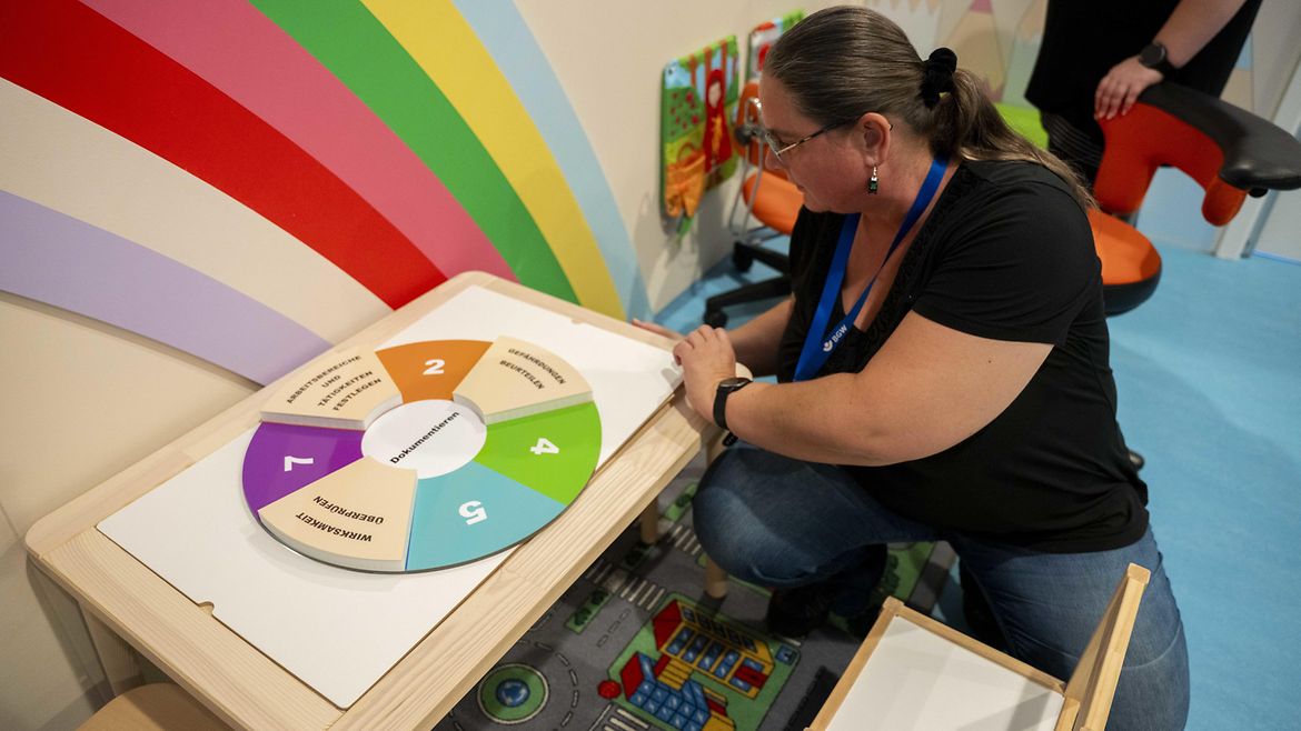 Eine Frau Kita kniet vor einem kleinen Spieltisch, auf dem ein Spiel zum Thema "Gefährdungsbeurteilung" liegt. Das Spiel hat die Form eines Glücksrades, in der Mitte steht "Dokumentieren", drei Felder sind bereits belegt. "Arbeitsbereiche und Tätigkeiten festlegen" (Schritt 1), "Gefährdungen beurteilen (Schritt 3) und "Wirksamkeit überprüfen" (Schritt 6).