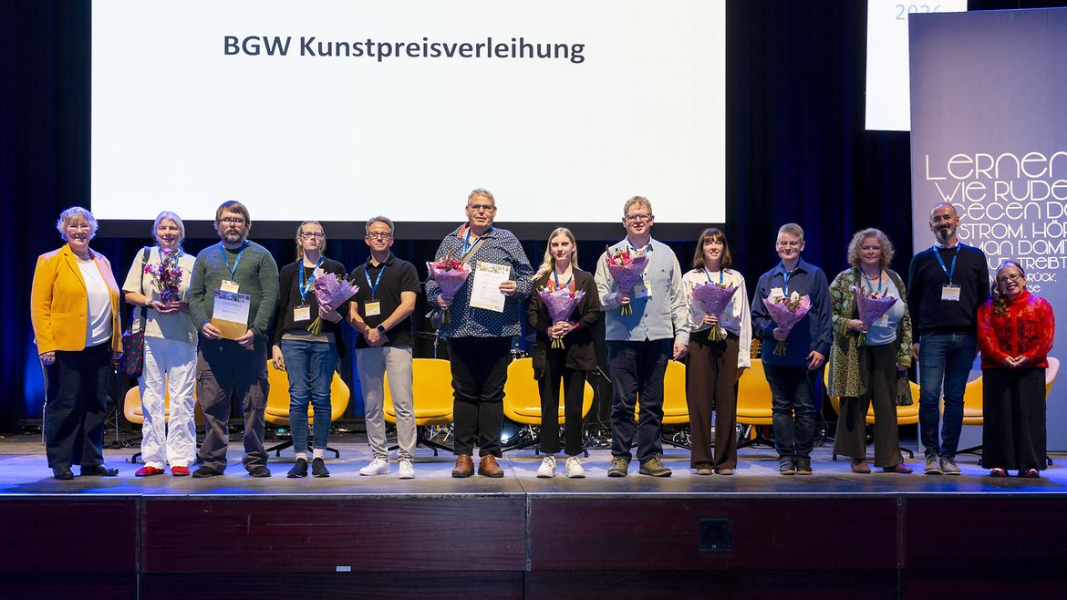 Dreizehn Personen stehen in einer Reihe auf einer Bühne, einige halten Blumen, andere Urkunden in den Händen. Im Hintergrund auf einer leuchtenden Leinwand steht "BGW Kunstpreisverleihung"