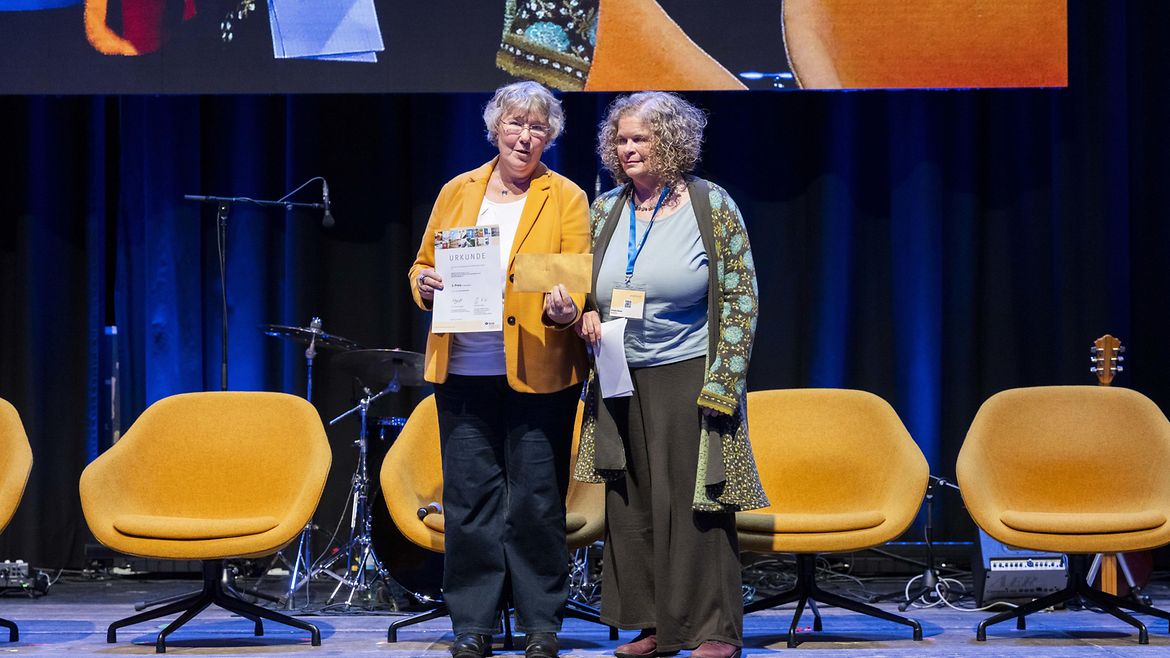 Silvia Thimm (l.) aus der BGW Selbstverwaltung mit der Gewinnerin des 3. Platzes Ursula Ebner auf einer Bühne.