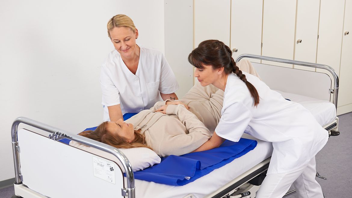 Kleine Hilfsmittel gegen Rückenbeschwerden: Gleitmatten Zwei Pflegekräfte lagern eine Patientin im Krankenbett mithilfe einer Gleitmatte.