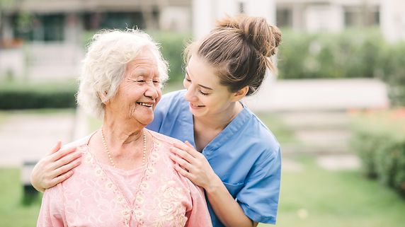 Lächelnde junge Pflegerin mit Seniorin im Freien.