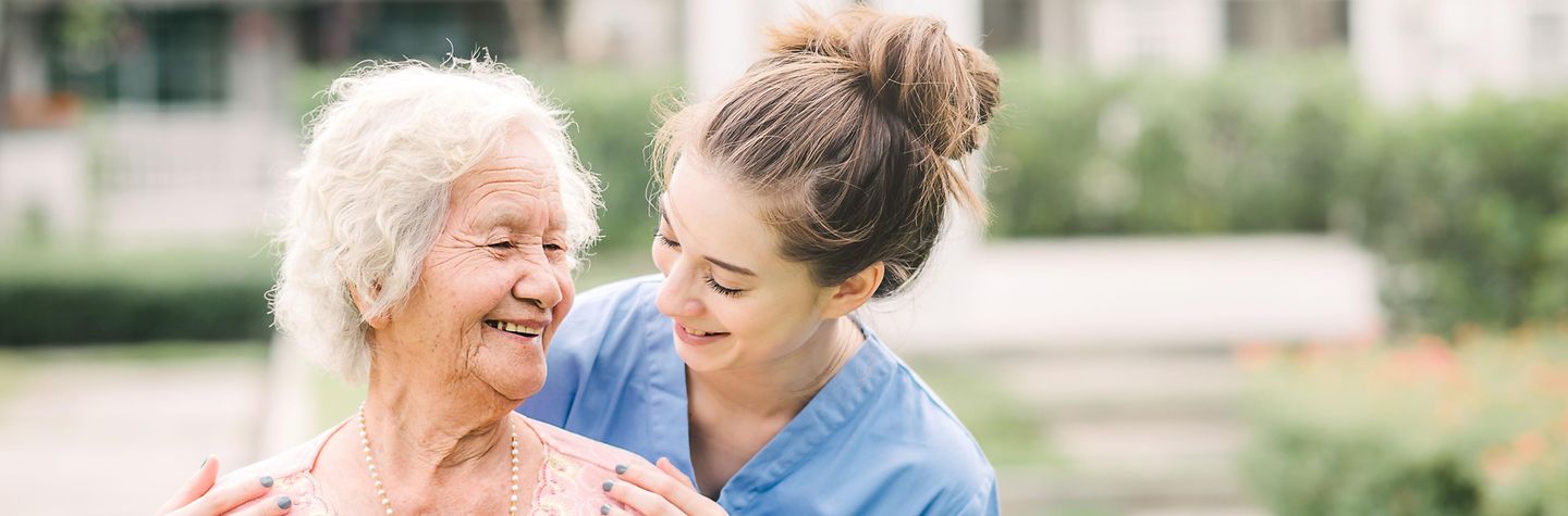 Lächelnde junge Pflegerin mit Seniorin im Freien.