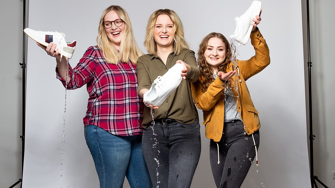 Drei junge Frauen vor einer weißen Wand mit Schuhen in der Hand, aus denen Wasser läuft.