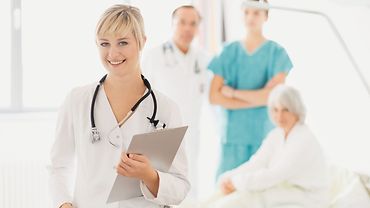 Im Vordergrund eine Ärztin mit Stethoskop und Klemmbrett. Im Hintergrund stehen ein Pfleger und ein Arzt am Bett eines Patienten.
