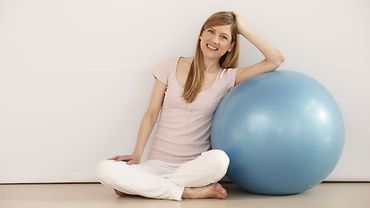 Junge Frau sitzt auf dem Boden und stützt sich an einem Gymnastikball ab.