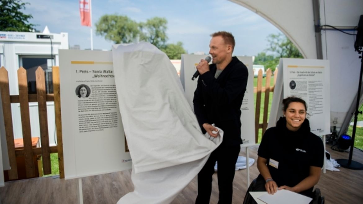 Mann mit Mikro in der Hand und Frau im Rollstuhl stehen vor einer Schautafel mit dem erstplatzierten Bild. Mann entfernt ein weißes Tuch von der Tafel.