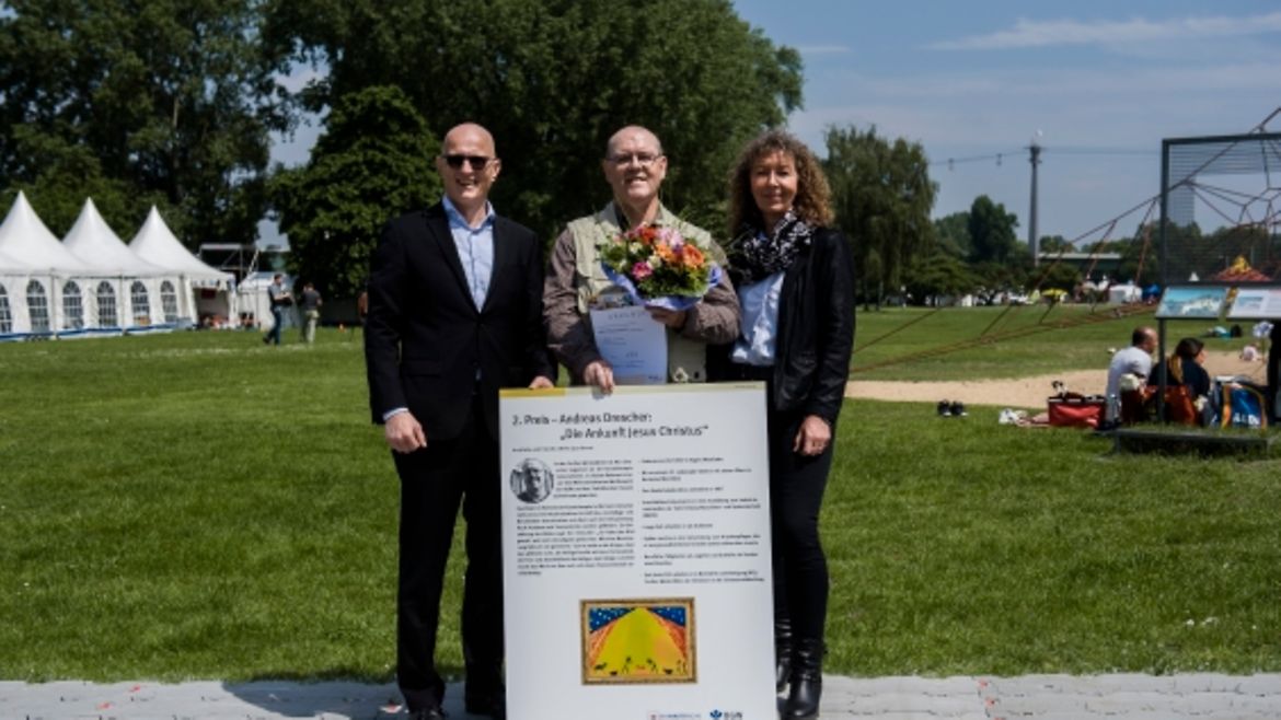 Von links: Jörg Schudmann (BGW), Andreas Drescher mit einem Plakat, auf dem das Bild "Die Ankunft Jesus Christus" zu sehen ist.