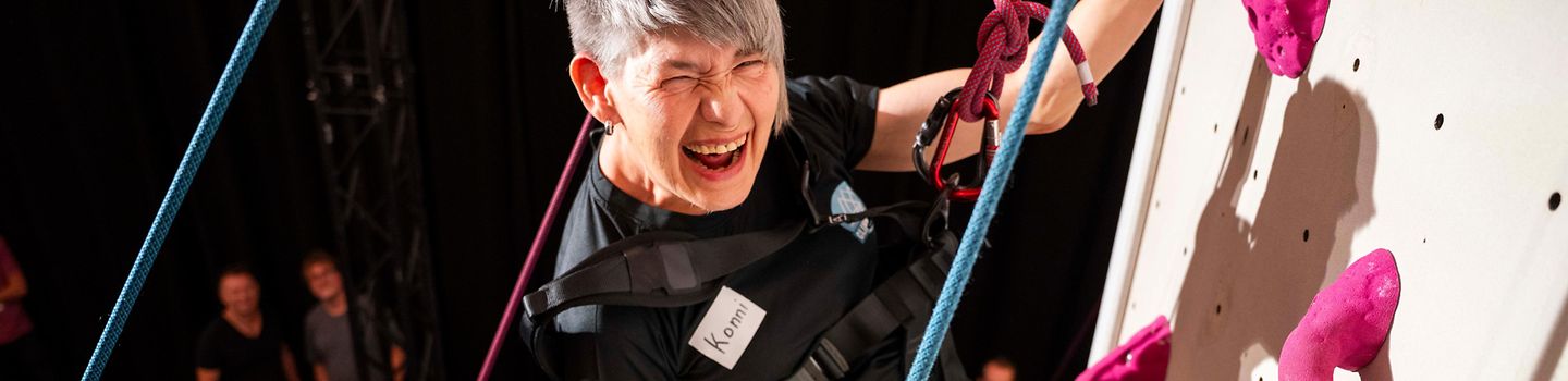Eine Frau hängt im Klettergurt am Kletterturm und lacht, im Hintergrund Zuschauende.