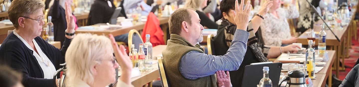 Selbstverwaltungsmitglieder an Reihen von Konferenztischen in einem Saal. Einige heben für die Abstimmung eine Hand.
