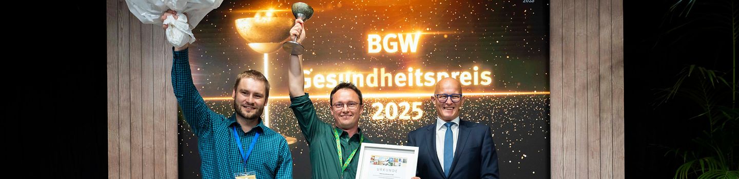 Die beiden Gewinner des Gesundheitspreises mit Blumenstrauß und Preis in der Hand auf einer Bühne neben dem BGW-Hauptgeschäftsführer