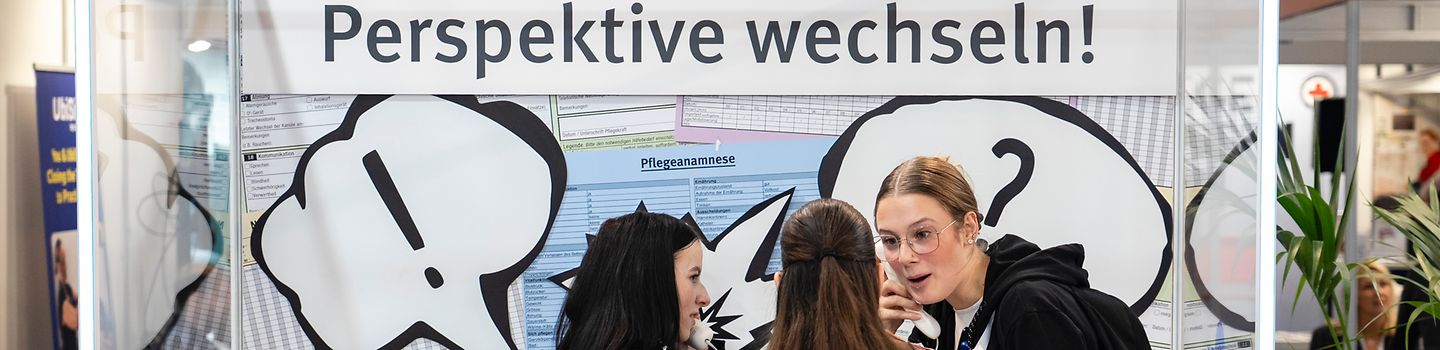 Drei junge Frauen stehen in einer Box eines Messestands und diskutieren. Hinter ihnen die Aufschrift "Konfliktbox?! Perspektive wechseln!"