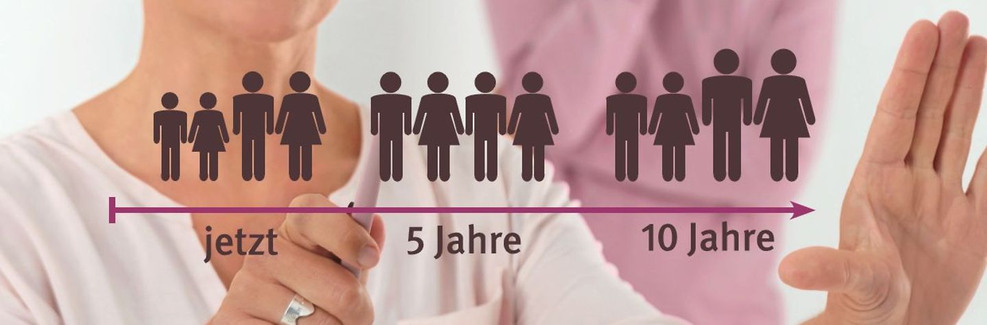 Frau deutet auf eine Grafik mit Figuren, die unterschiedliche Altersgruppen symbolisieren. Im Hintergrund ist unscharf ein junger Kollege zu sehen