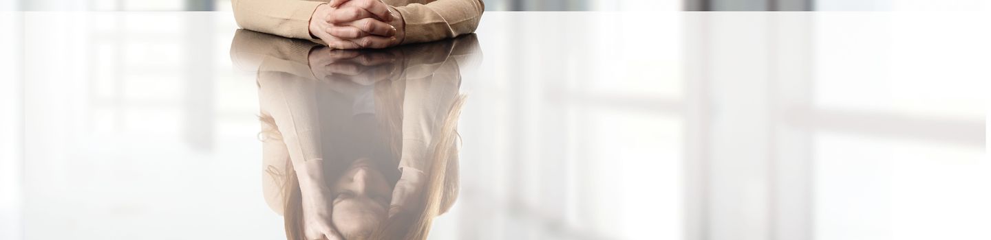 Eine Frau sitzt mit gefalteten Händen an einem Tisch in einem leeren Raum.