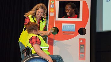 Aus der Kompetent mobil Show: Akteurin zeigt Rollstuhlnutzerin die Bedienung eines Fahrkartenautomaten