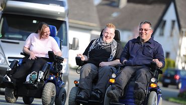 Eine Rollstuhlfahrerin und zwei Rollstuhlfahrer auf der Straße. 