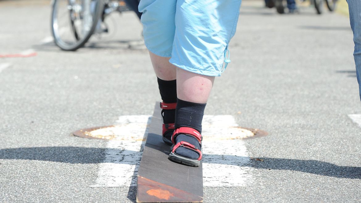 Junge Frau balanciert über einen Balken. Eine andere Person hält dabei ihre Hand.