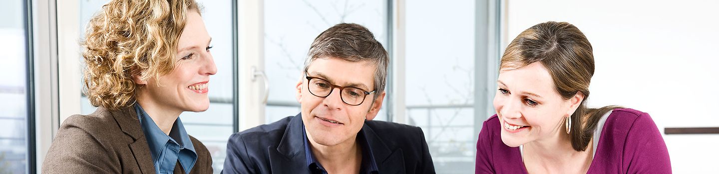 Gesundheitsmanagement Zwei Frauen und ein Mann im Gespräch an einem Konferenztisch. Die Kleidung ist legerer Businessstil.