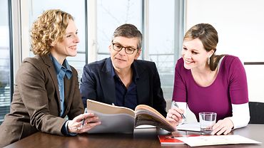 Zwei Frauen und ein Mann im Gespräch an einem Konferenztisch. Die Kleidung ist legerer Businessstil.