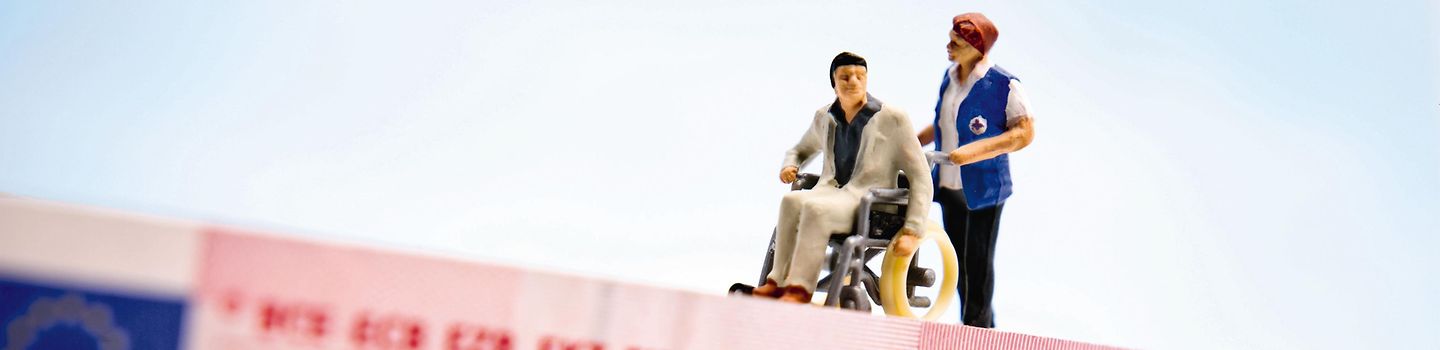 Figuren und Geldschein Kleine Figur eines Rollstuhlfahrers wird von der Figur einer Helferin auf 10-Euro-Schein geschoben