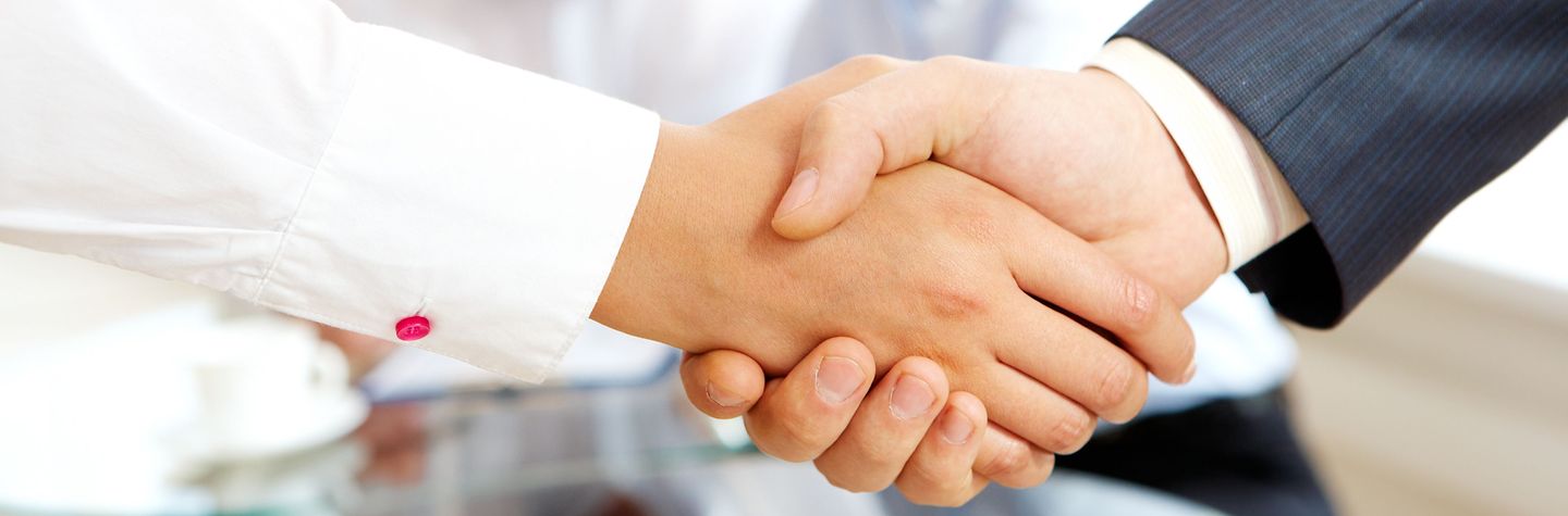 Handschlag Nahaufnahme Händeschütteln, im Bildhintergrund unterhalten sich zwei Personen