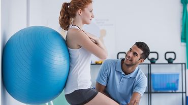 Junge Frau macht eine Übung mit einem Gymnastikball. Ein Physiotherapeut kontrolliert die Übung.