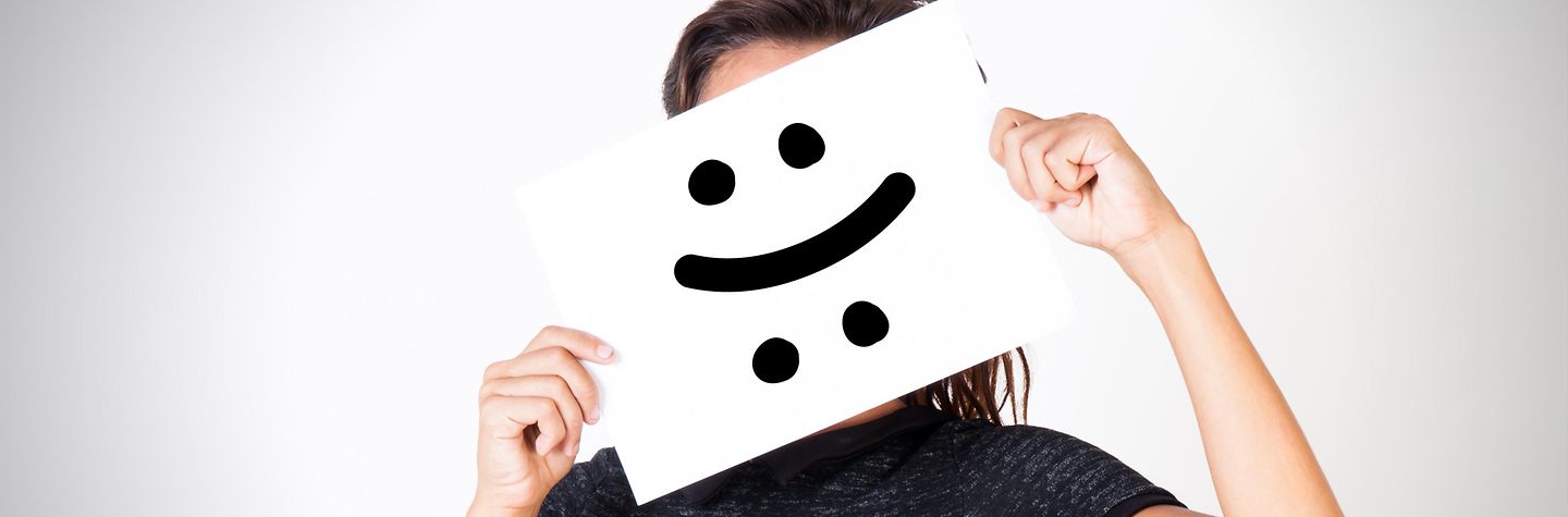 Eine Person hält ein Bild mit gut und schlechtgelauntem Smiley vors Gesicht.