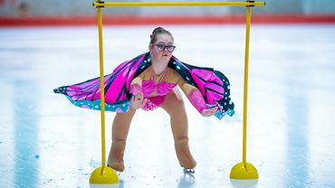 Junge Eislaufkünstlerin mit pinkem Umhang fährt unter Hindernis hindurch
