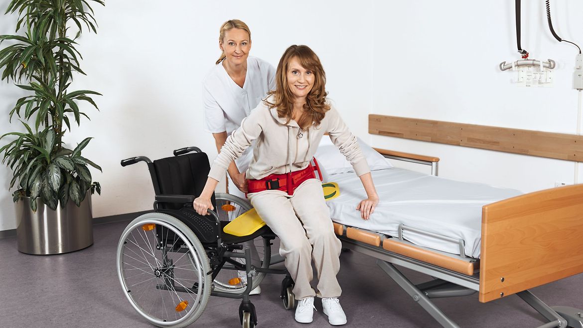 Pflegerin hilft einer Patientin mithilfe eines Rutschbretts und eines Haltegürtels vom Bett in den Rollstuhl