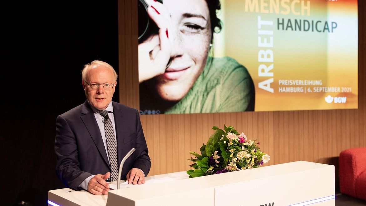 Prof. Dr. Stephan Brandenburg am Rednerpult, im Hintergrund Einblendung des Logos "Mensch - Arbeit - Handicap" und Bild einer Frau mit Kamera